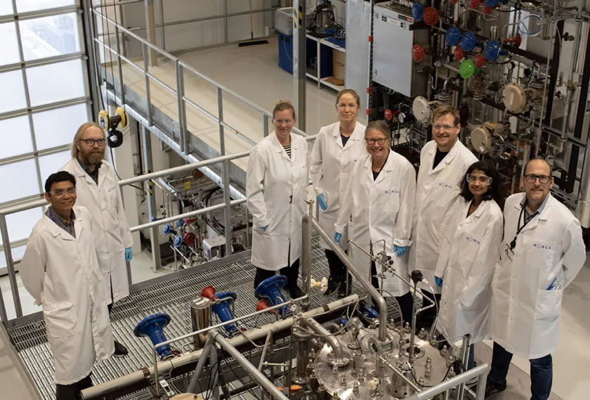 Åtte personer med labfrakk i laboratorieomgivelser.